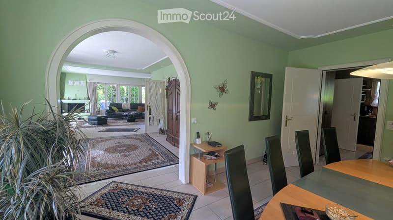 A well-lit living and dining area with light green walls, tiled flooring, and large windows. The living area features a sofa, coffee table, ottoman, and a small side table with a lamp. The dining area includes a wooden table with chairs and a decorative b