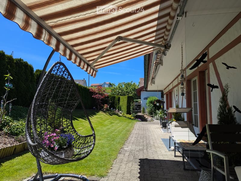 Garden with a covered patio, wicker hanging egg chair with flowers, lounge chairs, and a well-maintained lawn.