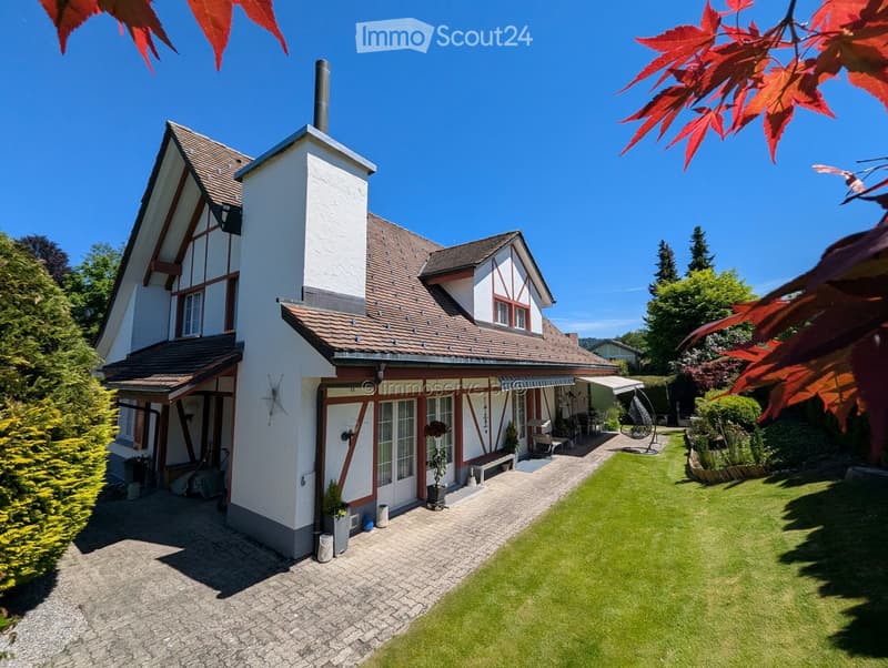 Two-story house with red-tiled roof, white and red facade, chimney, and well-maintained garden with lawn, patio, and outdoor seating area.