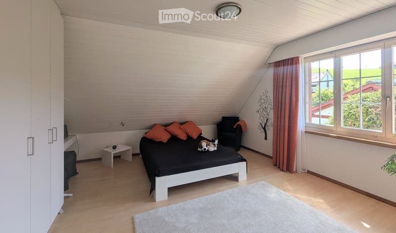 Bright bedroom with wooden flooring, large windows, a white bed with orange pillows, a nightstand, a small chair, and a decorative wall art piece.