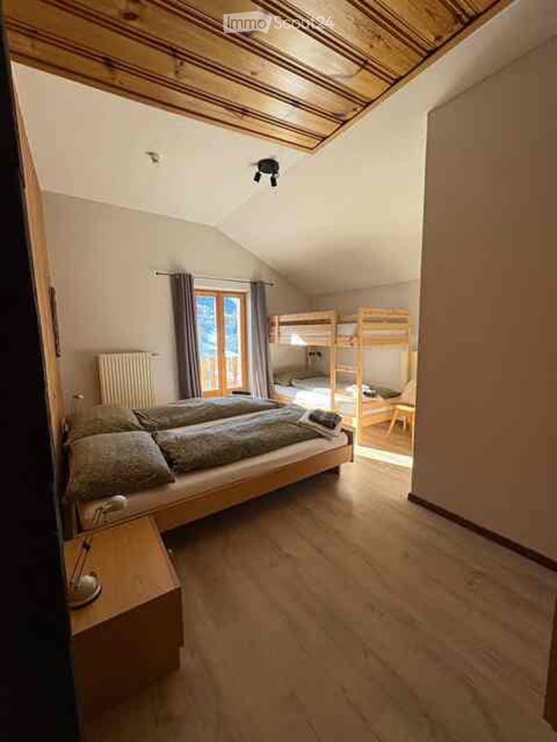 Cozy attic bedroom with wooden floors, slanted wooden ceiling, large window with view, double bed with pillows and blankets, nightstand, radiator, and bunk bed in the background.