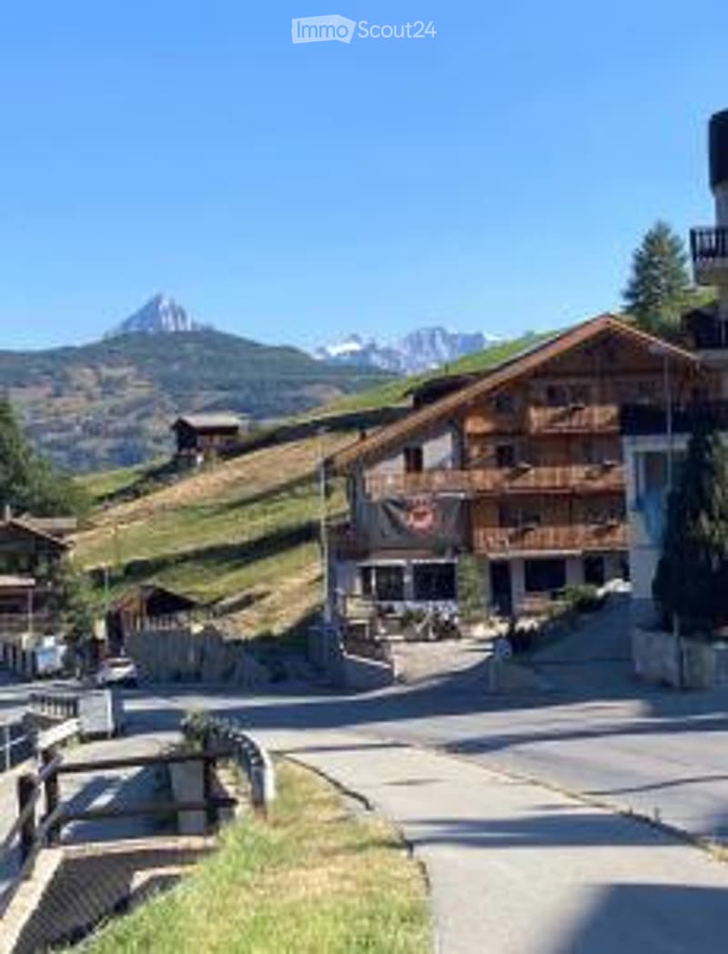 Traditional wooden chalet-style building in a picturesque mountain village, surrounded by greenery and snow-capped peaks.