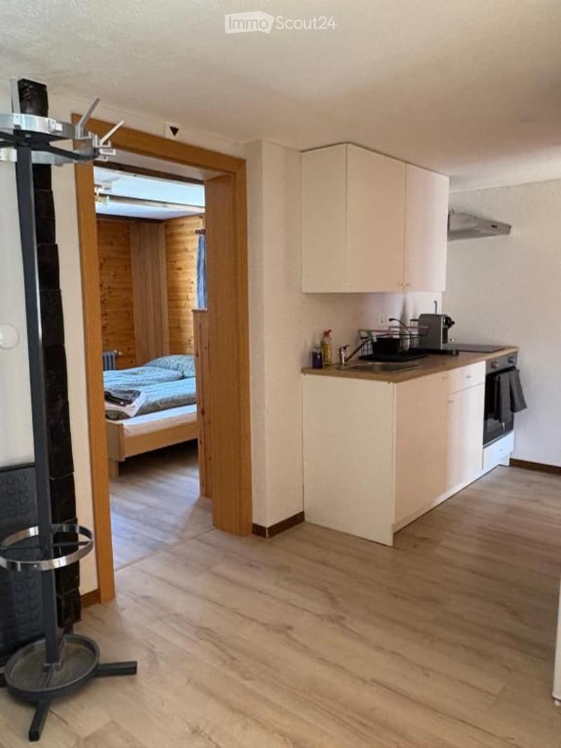 A modern, compact kitchen with white cabinets, a sink, stove, and oven. A small bedroom is visible through an open doorway, featuring a bed with pillows and a blanket.
