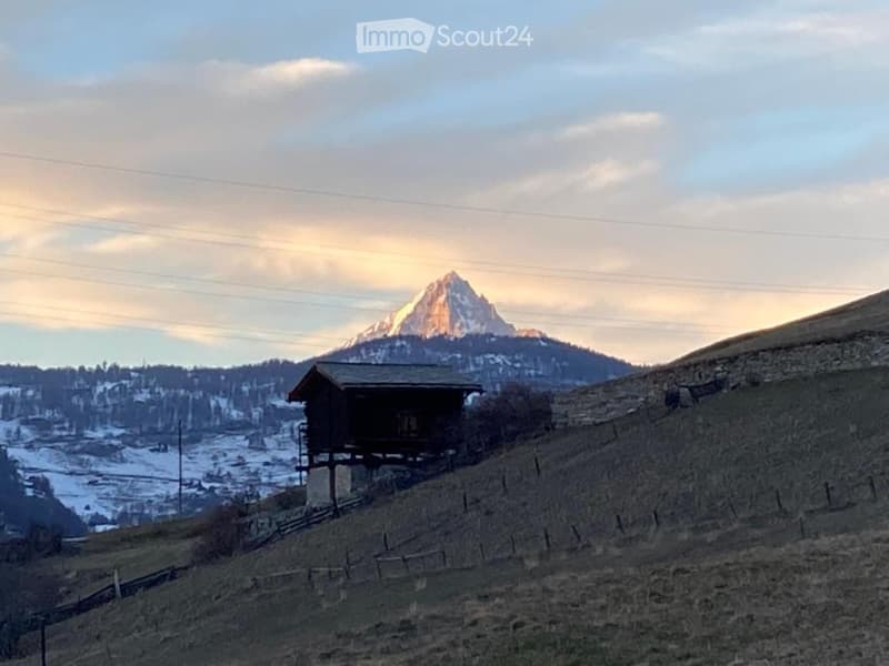 A wooden house sits on stilts in a hilly, snow-covered landscape with a mountain peak in the background.
