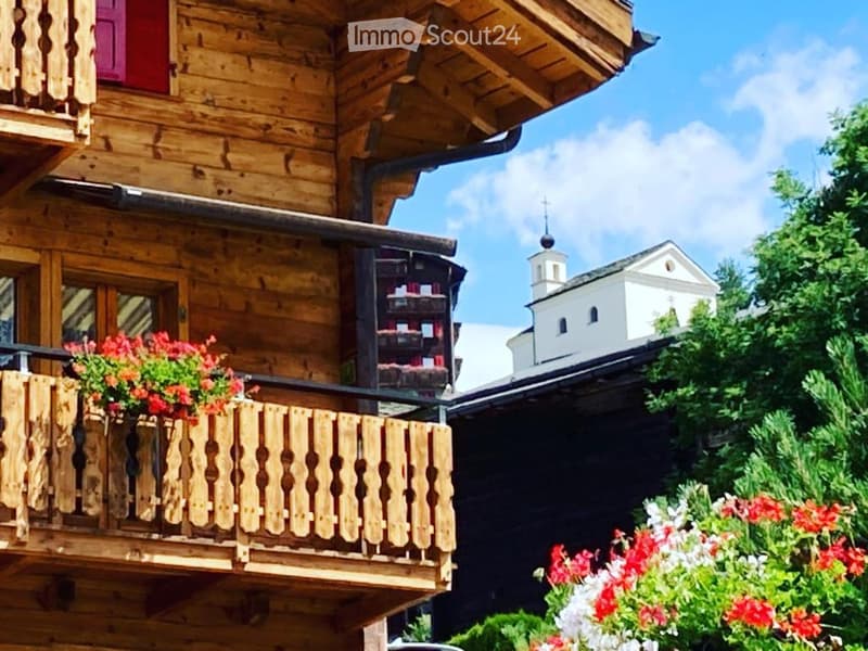A wooden chalet with a balcony decorated with red and white flowers, overlooking a village with a church in the background.
