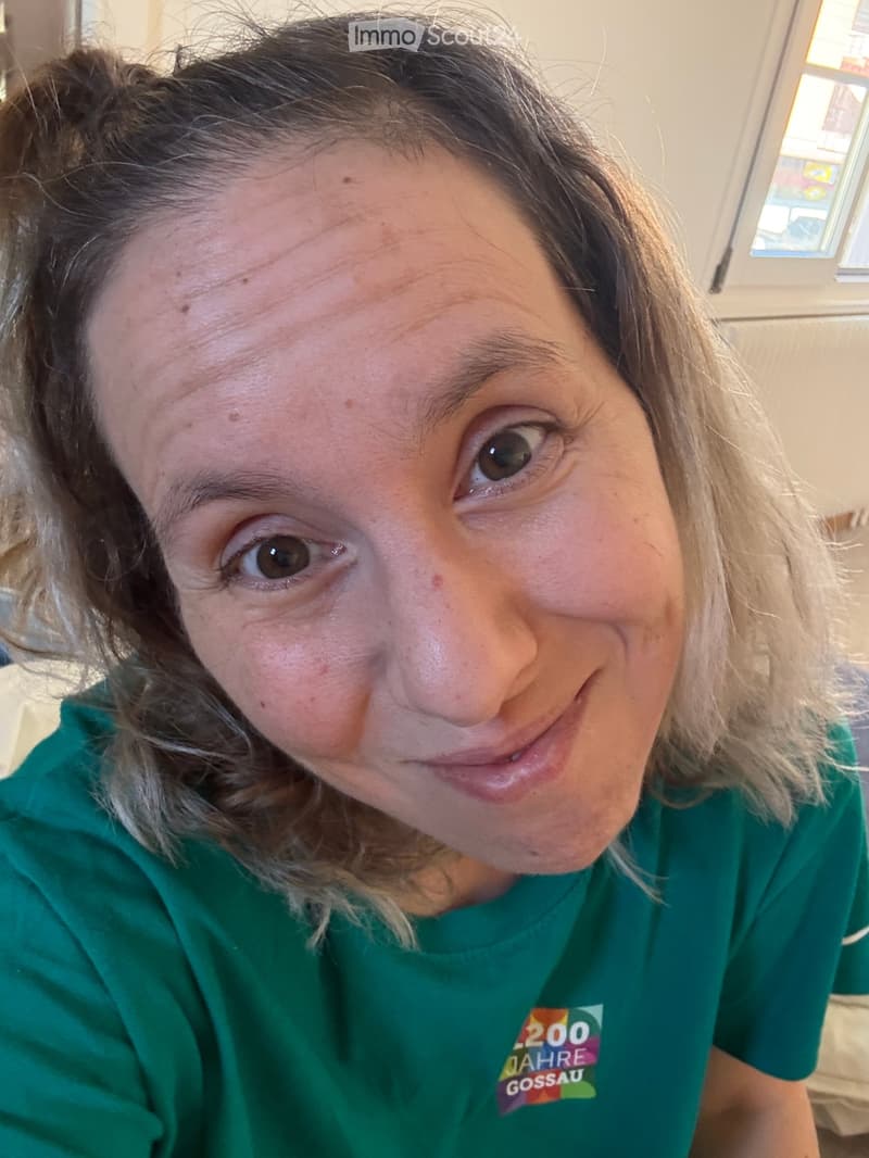 A person with long, light brown hair is smiling at the camera, wearing a green t-shirt with a logo that reads "200 JAHRE GOSSAU". The background shows a window and part of a room with light-colored walls.