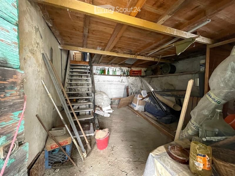 An unfinished attic space with exposed wooden beams, a metal staircase leading up, and various stored items including tools, containers, and bags.