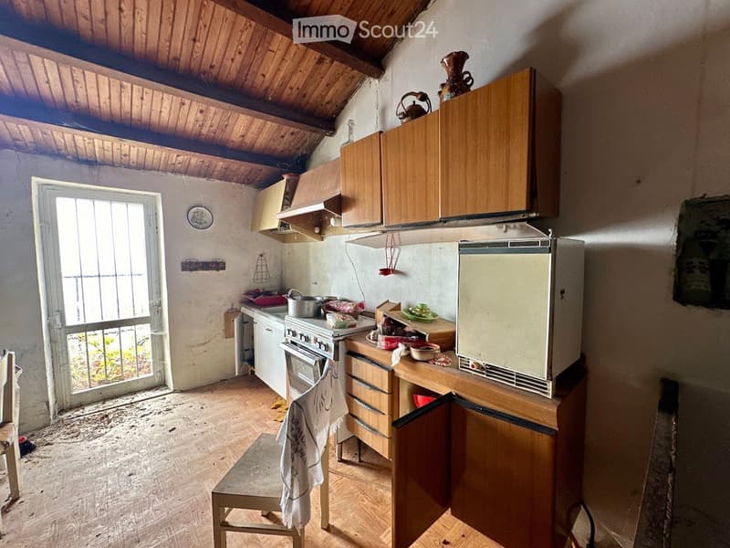 Small kitchen with wooden cabinets, a refrigerator, an oven, and a countertop with various items. The room has a slanted wooden ceiling and a window with bars.