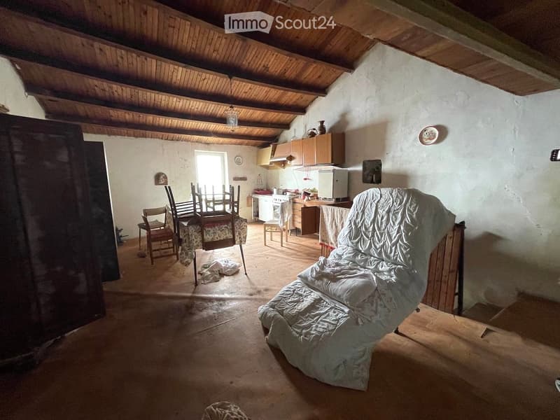 A rustic, open-plan living and dining area with wooden beams, a covered sofa, a dining table with chairs, and a small kitchen area with cabinets and appliances.