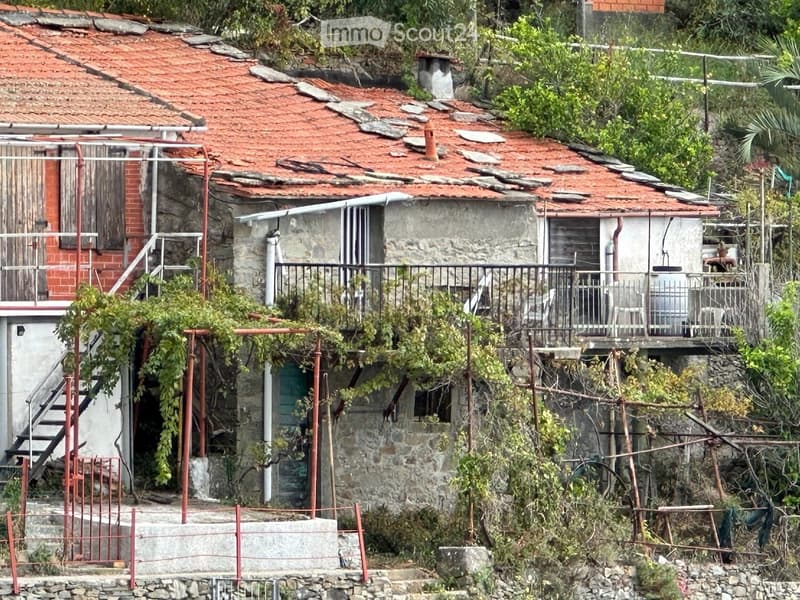 A rustic stone house with a red-tiled roof, surrounded by greenery and a small balcony with metal railings.