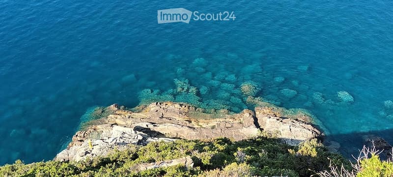 A scenic coastal view with clear blue water, rocky outcrops, and sparse vegetation on a cliffside.