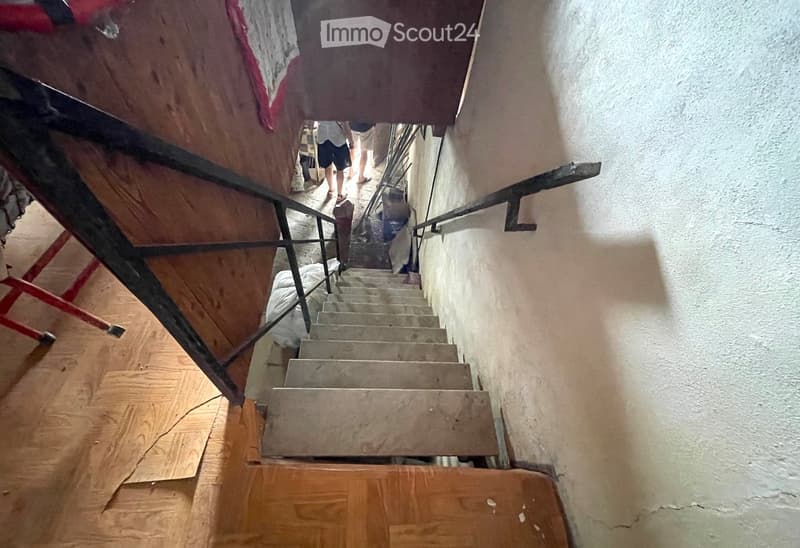 A narrow staircase with wooden steps and metal railings, leading upwards. The walls are plain and white, and the floor is covered with wooden planks. There are some people visible in the background, and a red ladder is partially visible on the left side.