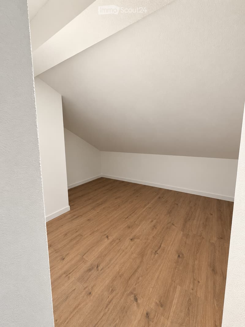 An empty room with light wooden flooring and white walls, featuring a slanted ceiling typical of an attic space.