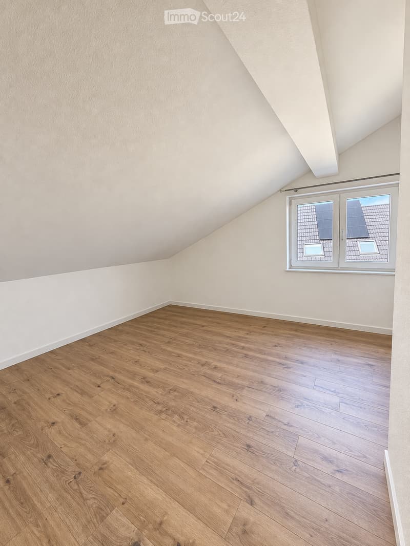 Empty attic room with wooden flooring and white walls, featuring a large window with a view of the roof.
