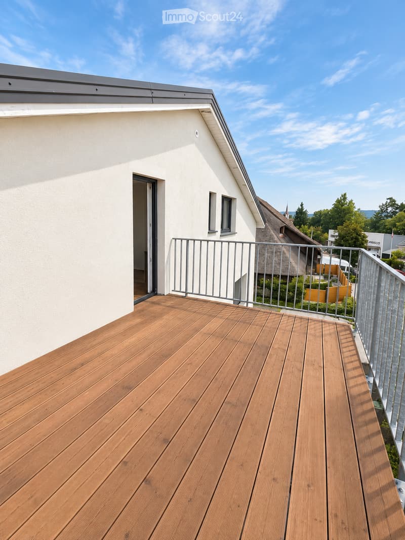 Wooden deck with metal railing, open to the sky, with a view of a house and surrounding greenery.