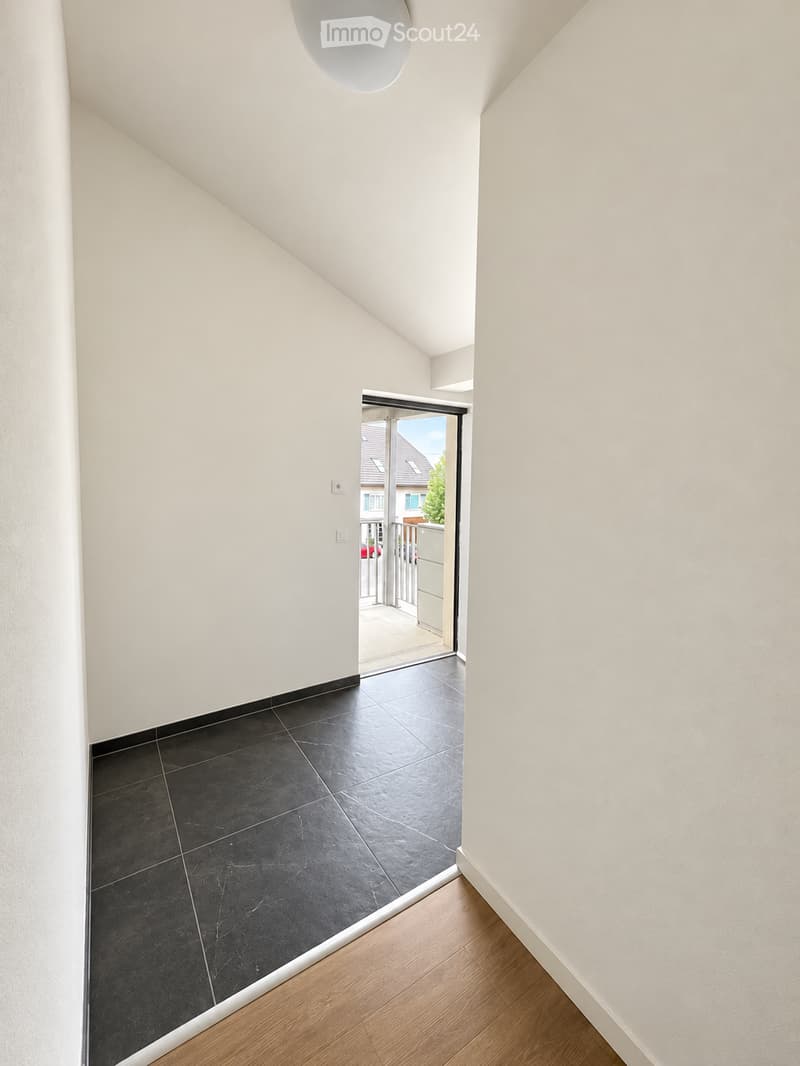 Modern hallway with white walls, dark tiled floor, and a large skylight above. An open doorway leads to a balcony area with outdoor tiles and a view of neighboring houses.