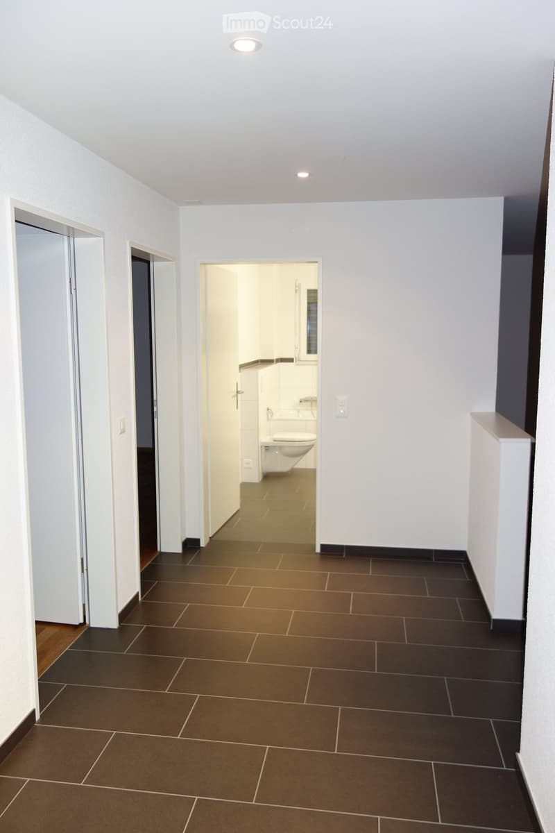 Empty hallway with tiled floor, white walls, and recessed lighting. A partially open door leads to a bathroom with a toilet and sink.