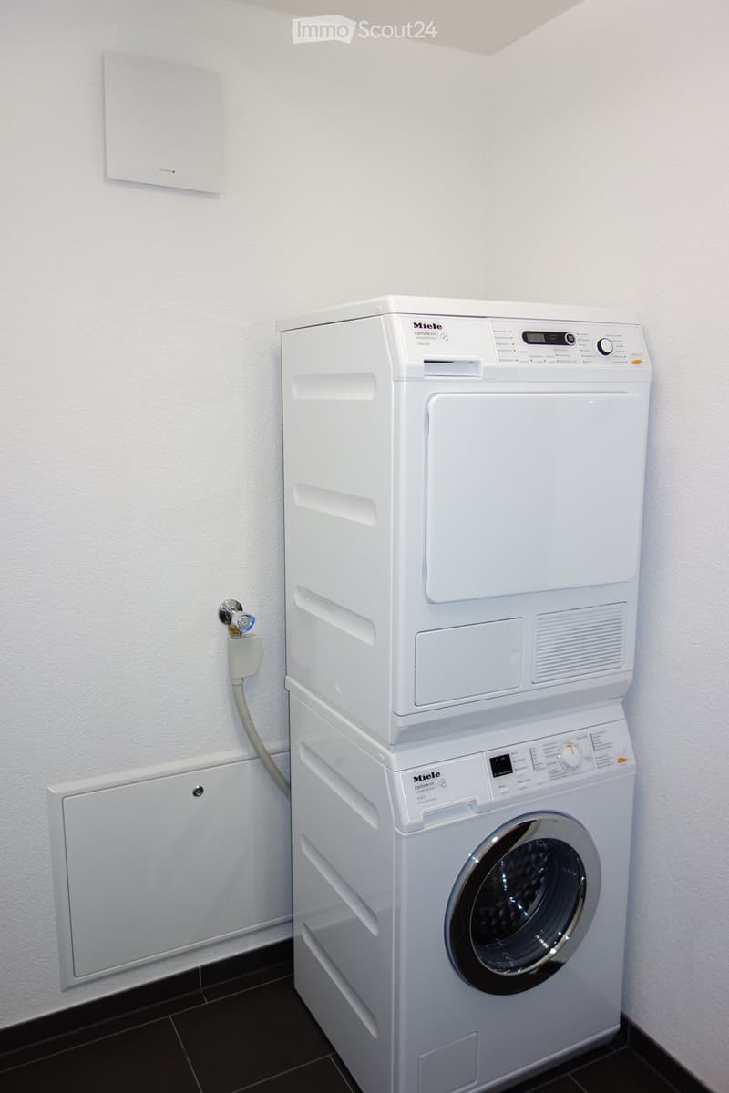 A compact bathroom with a stacked white washing machine and dryer unit, featuring a control panel and a water connection.