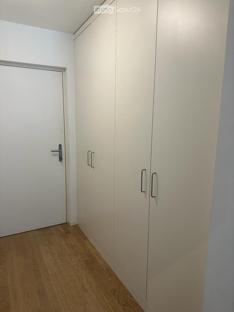 Empty room with light wooden flooring, white walls, and built-in wardrobes with silver handles. A closed door is visible on the left side.