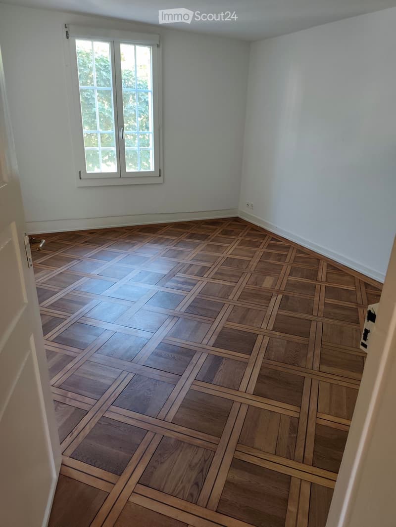Empty room with light-colored walls and a herringbone-patterned wooden floor. A large window with white frames is visible on one wall.
