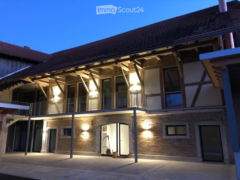 Two-story building with exposed wooden beams, brick walls, and a balcony with metal railing. The entrance features a large glass door and a small open area with a vacuum cleaner visible inside.