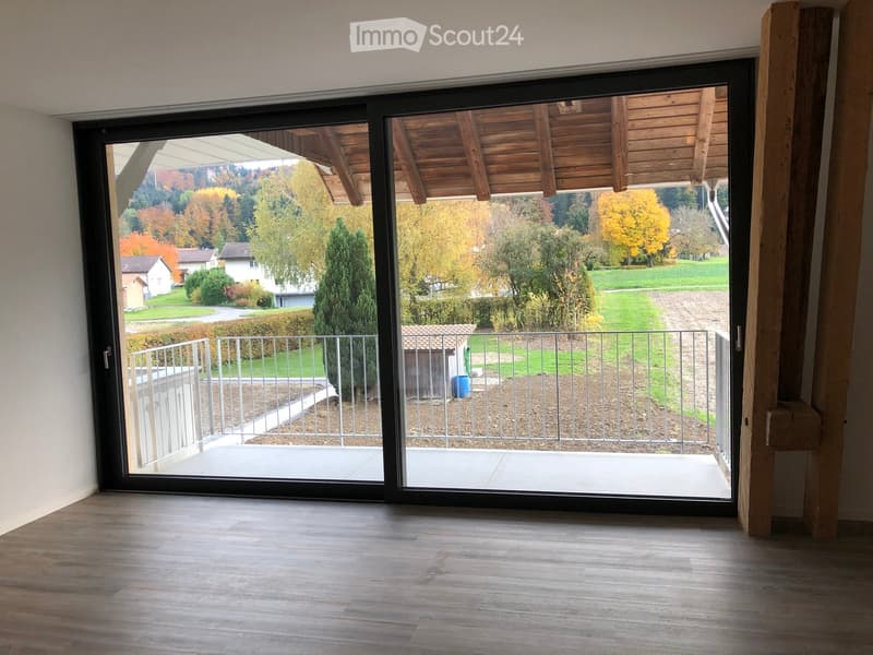 Empty room with wooden flooring and large sliding glass doors leading to a balcony with a wooden roof and metal railing, overlooking a scenic autumn landscape.