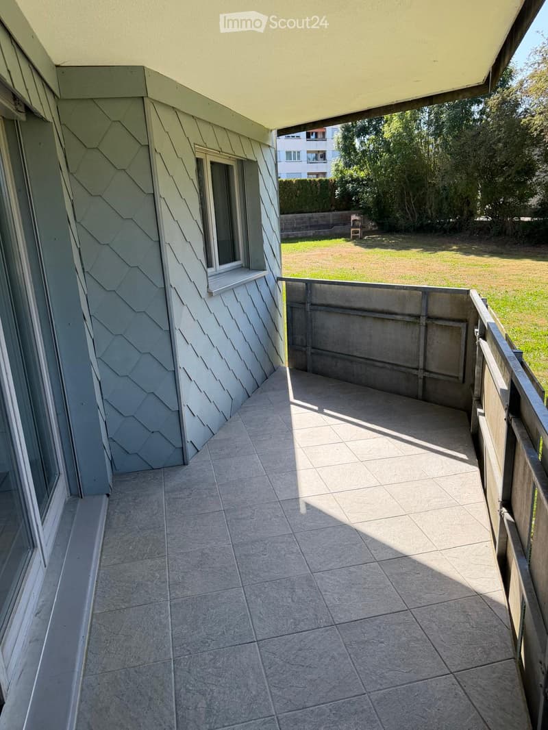 Balcony with tiled floor, metal railing, and a view of a grassy area with trees and a building in the background.