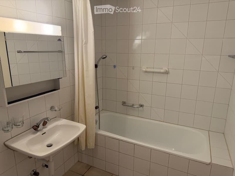 Bathroom with white tiled walls and floor, featuring a sink, bathtub, mirror, and two cups on a shelf.