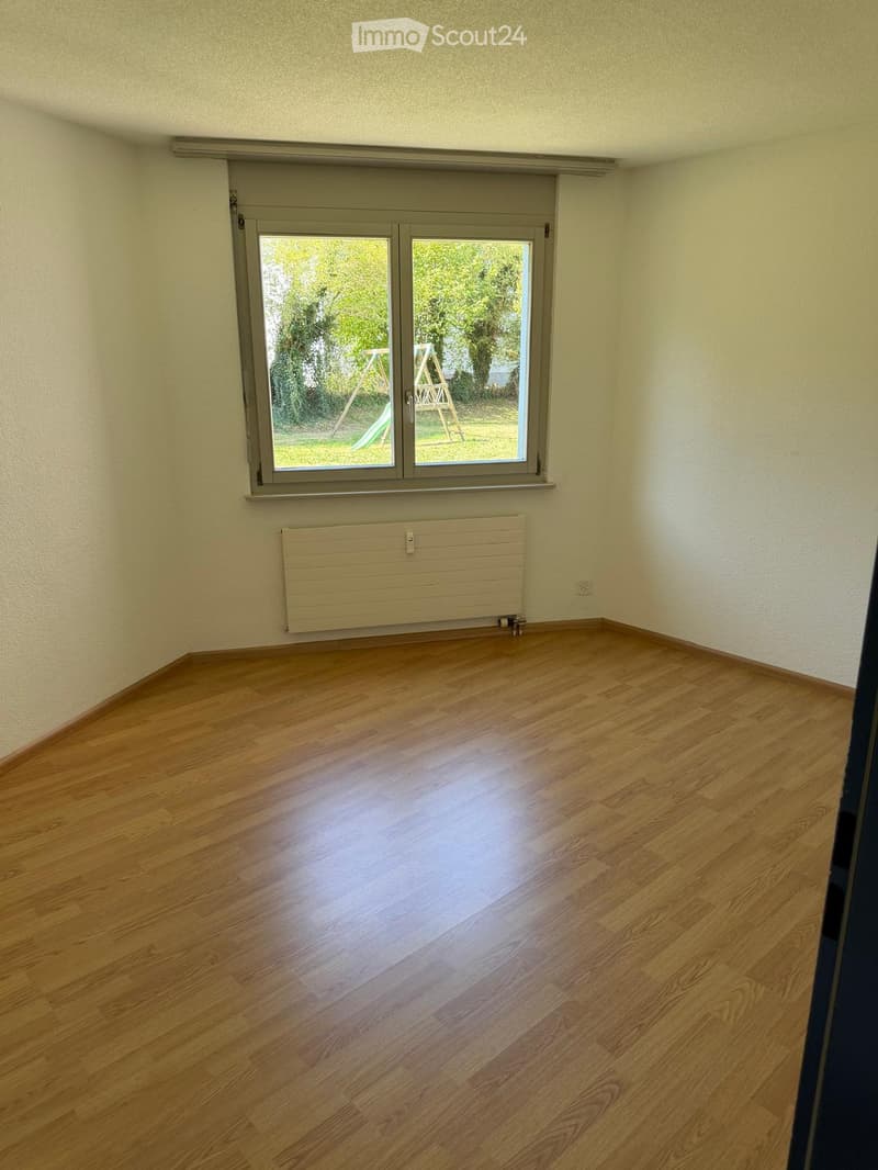 Empty room with wooden flooring, white walls, a large window with blinds, and a radiator below the window.