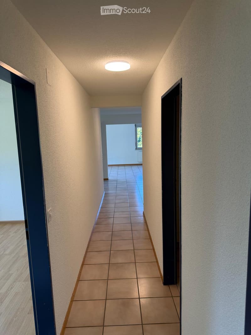 A narrow hallway with tiled flooring, beige walls, and a ceiling light. Doors are visible on both sides, leading to other rooms.