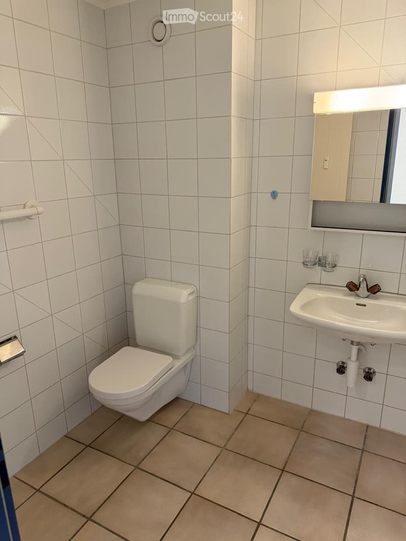 Modern bathroom with white tiled walls, beige tiled floor, toilet, sink, mirror, and two cups on the sink counter.