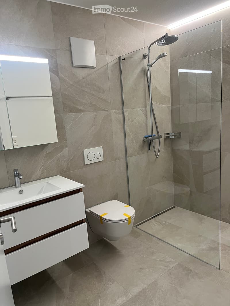 Modern bathroom with a white sink, mirror, toilet, and glass-enclosed shower with a handheld showerhead. The walls and floor are tiled in a light beige marble pattern.