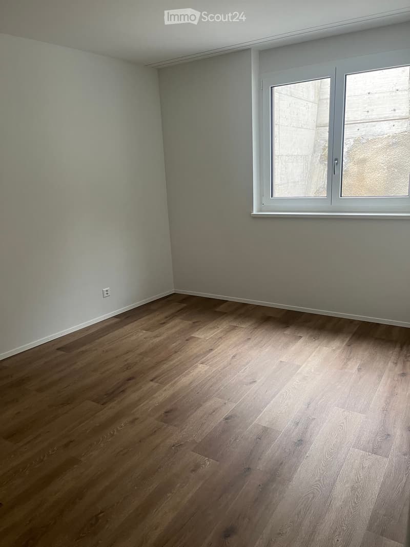 Empty room with wooden flooring and white walls, featuring a large window with white frame and a visible electrical outlet on the wall.