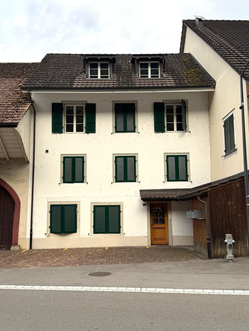 Two-story building with white walls, green shutters, and a wooden front door. The roof is tiled, and there is a small balcony area above the entrance.