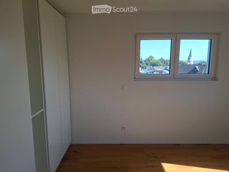 Empty room with wooden flooring, white walls, and a large window offering a view of a town with a church spire.