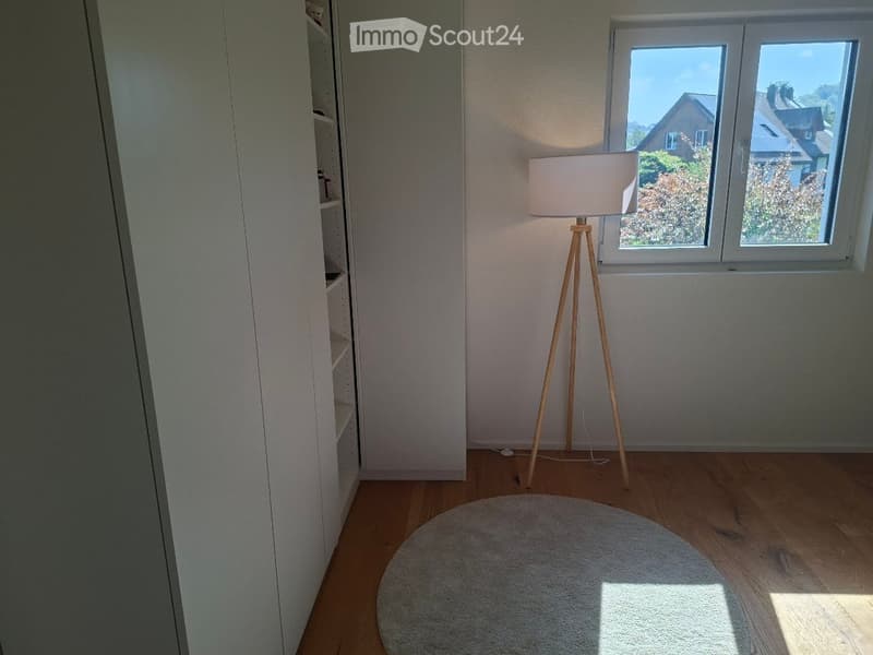 Empty room with wooden flooring, white walls, a round rug, a floor lamp, and a window with a view of the outside.