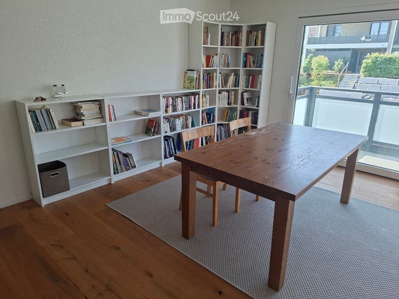 A well-lit room with wooden flooring, a large wooden table, two chairs, and multiple white bookshelves filled with books and decorative items. A large window with a view of a balcony and outdoor area is visible.