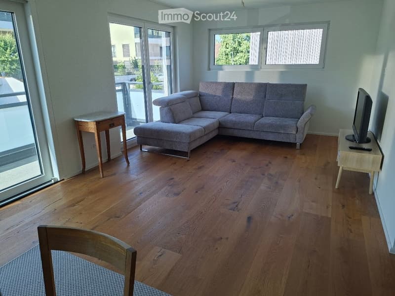 Modern living room with wooden flooring, large windows, and a grey sectional sofa. Includes a small wooden table, a television, and a wooden chair.