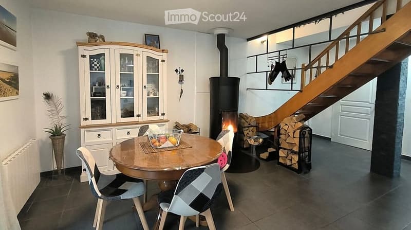 Bright dining area with a round wooden table, four chairs, a cabinet with glass doors, a fireplace, and a staircase leading to the upper floor.