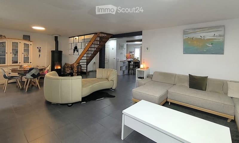 Modern living room with a curved sofa, coffee table, dining area with chairs and table, a cabinet, and a fireplace. The space is well-lit with large windows and a staircase leading to an upper level.
