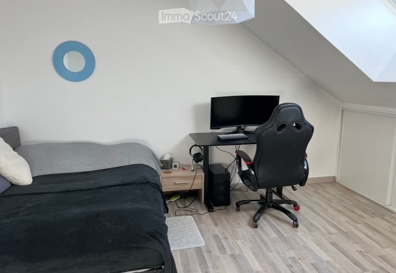 A cozy attic bedroom with a slanted ceiling, featuring a sofa, desk with a computer setup, and a gaming chair.
