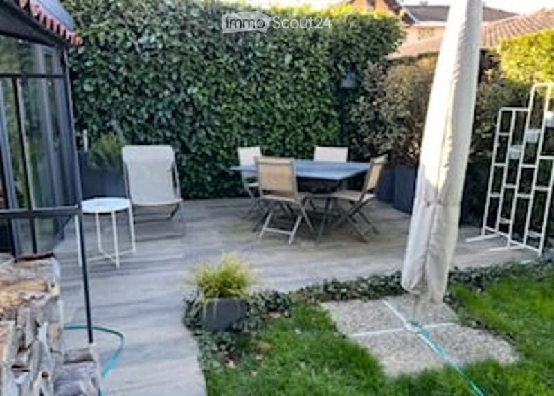 A small patio area with a dining set, including a table and chairs, surrounded by a lush green hedge and a stone wall. There is a small potted plant and a white umbrella nearby.