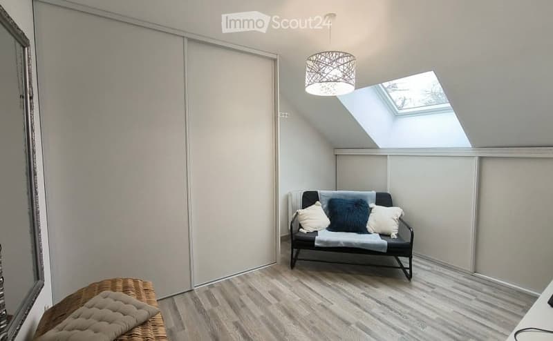 Bright, modern room with wooden flooring, a small sofa with decorative pillows, a wicker bench, and a skylight window allowing natural light.
