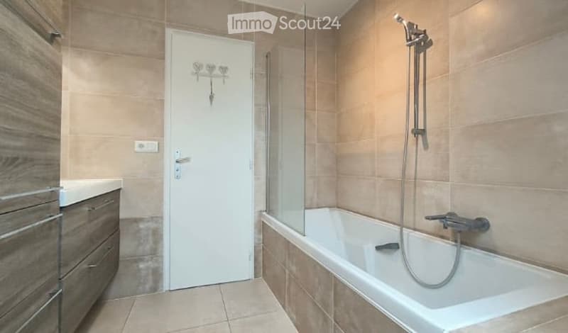 Modern bathroom with a bathtub, shower, and wooden cabinet. The walls are tiled in beige, and the floor is tiled in light beige.