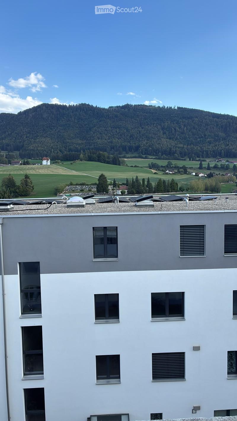 Modern two-story building with grey and white exterior, multiple windows, and solar panels on the roof, set against a scenic mountainous backdrop.