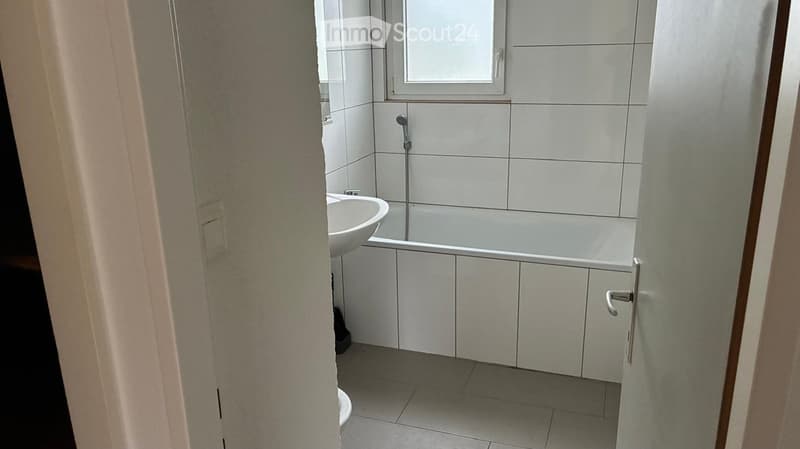 Modern bathroom with white tiles, a sink, a bathtub, and a cabinet. The space is clean and well-lit with a window providing natural light.