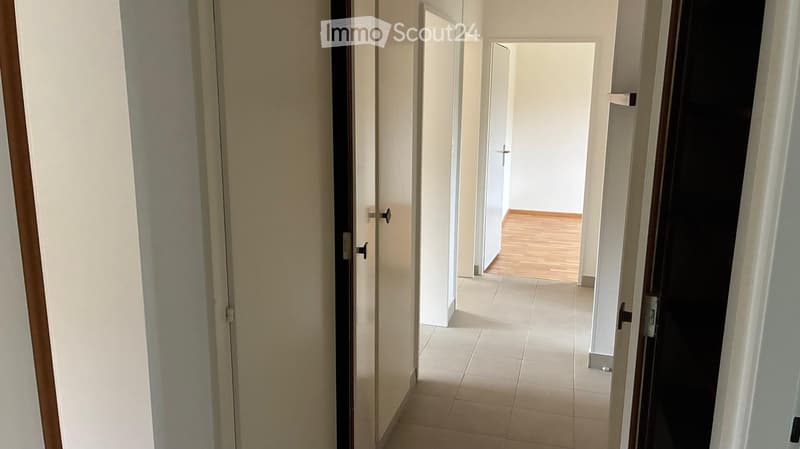 Empty hallway with tiled floor, wooden door, and open doorway leading to another room with wooden flooring.