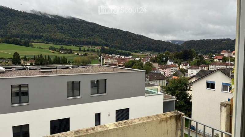 View from a balcony showing a modern building with grey and white facade, multiple windows, and a scenic valley with houses, green fields, and distant mountains.