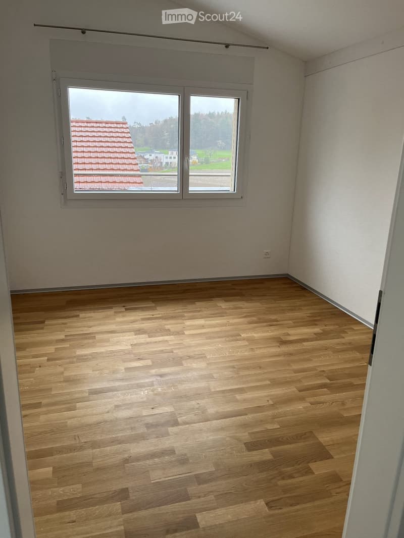 Empty room with wooden flooring and large window showing a view of a roofed building and greenery outside.