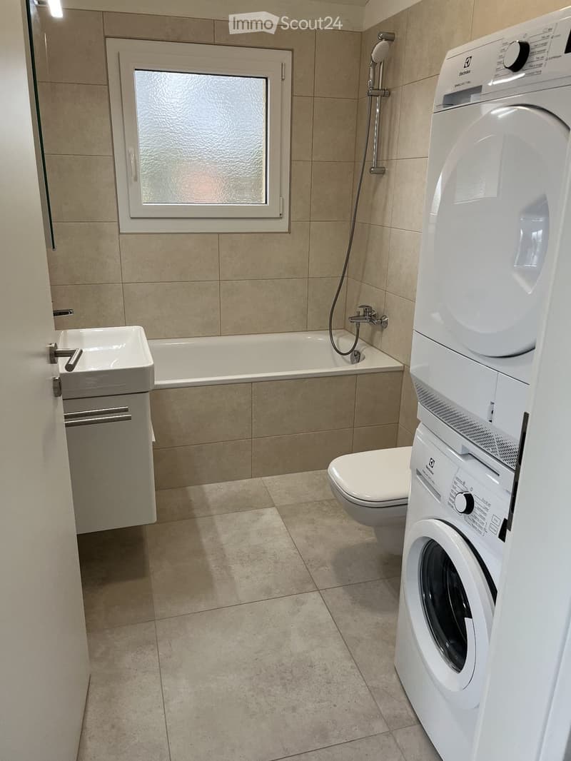 Modern bathroom with beige tiles, bathtub, toilet, sink, washing machine, and shower fixture.